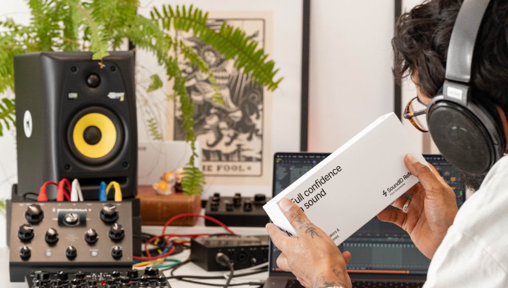 Music producer wearing headphones examines a SoundID Reference box in a modern home studio with speakers, synths, and plants.